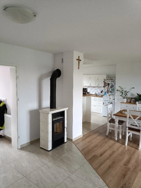 Wolnostojący piecyk kominkowy w białej kaflowej obudowie, zamontowany w jasnym, nowoczesnym wnętrzu domu. Piecyk stoi przy ścianie oddzielającej salon od kuchni, prezentując estetyczne i funkcjonalne wykonanie instalacji.